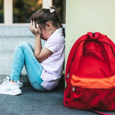 young girl with back up against wall in school yard with red backpack on ground